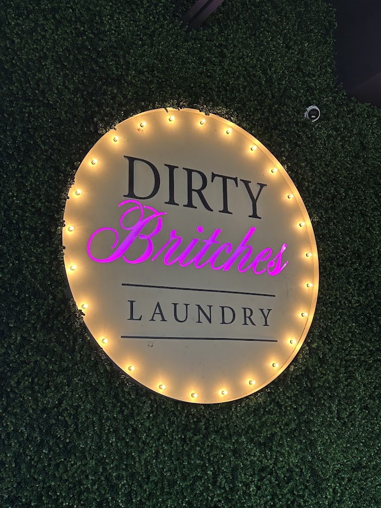 Marquee bulb-lit Dirty Britches Laundry sign on a green hedge wall